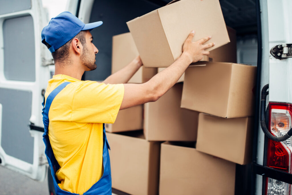 Cargo delivery service, male courier in uniform with box in hand unloads truck with cardboard parcels. Empty container